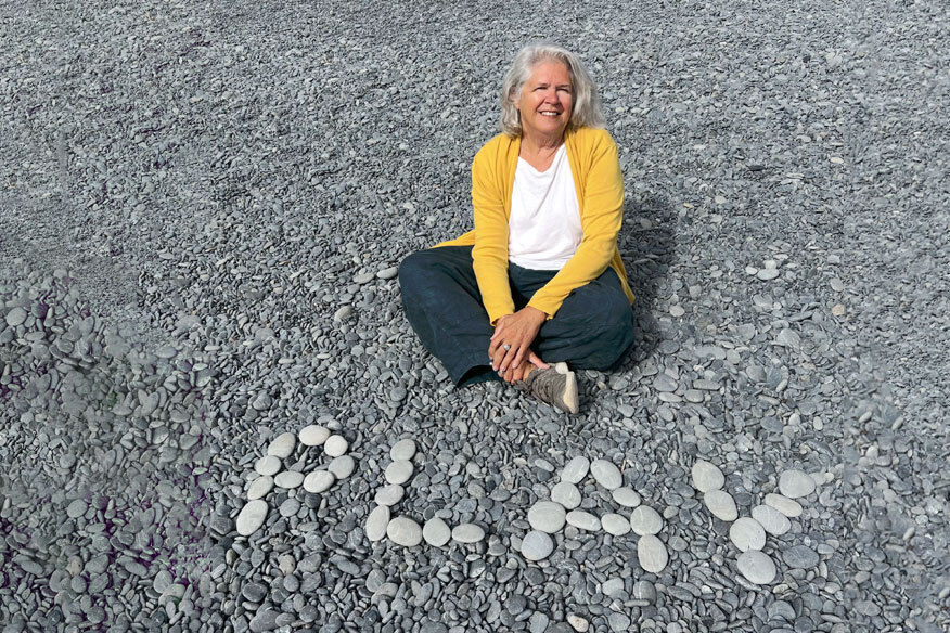 Virginia Hamilton sits on a pebble beach with rocks that spell PLAY